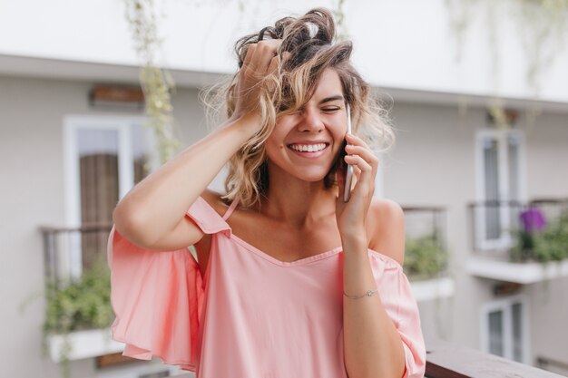 Close-up portrait of carefree blonde lady smiling and talking on phone. Outdoor photo of fascinating female model playing with her hair during call.