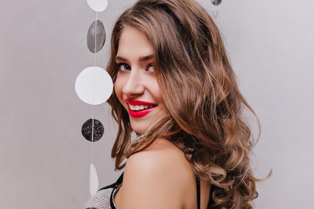 赤い口紅が肩越しに見ているのんきな巻き毛の女の子のクローズアップの肖像画。暗い壁に笑みを浮かべてロマンチックな髪型を持つ官能的な白人女性モデル。