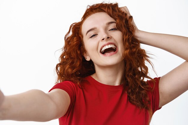 Close up portrait of carefree and happy ginger girl, touching her curly natural hair and laughing while taking selfie on white wall