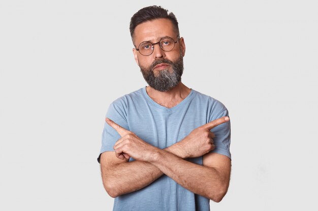 Close up portrait of Caucasian male points at different sides with index fingers, cant choose between two items, has calm expression, wearing glasses and gray t shirt, isolated over white wall.