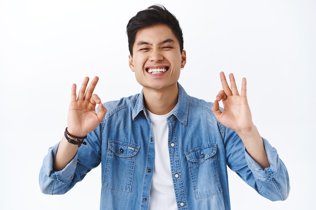 Close-up portrait of charismatic, attractive asian man guarantee best quality, recommend product, laughing and smiling pleased, being assertive you will like it, white wall.