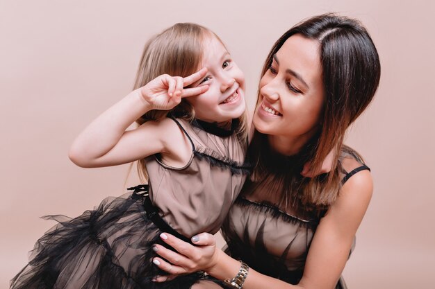 Close-up portrait of charming cute girl and her stylish mother wearing similar black dresses on beige wall