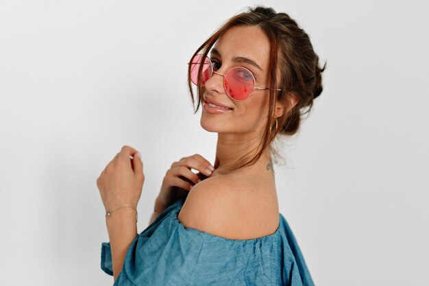 Крупным планом портрет очаровательной европейской загорелой девушки в круглых розовых очках, позирующей перед камерой сзади с милой улыбкой на изолированном фоне