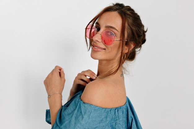 Close up portrait of charming european tanned girl in round pink glasses posing at camera from back with lovely smile over isolated background