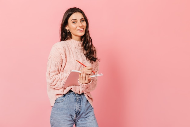 Close-up portrait of charming female student writing in her notebook. Lady in mom's jeans and sweater cute smiles on pink background.