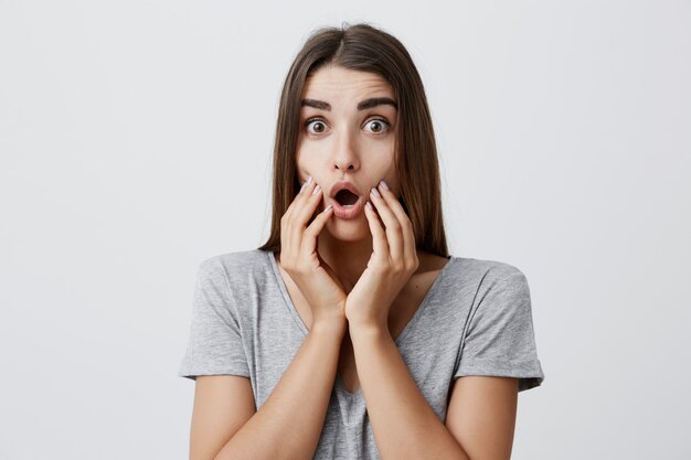 Close up portrait of charming good-looking joyful caucasian woman with long dark hair in casual stylish clothes holding hands on face with opened mouth and surprised expression