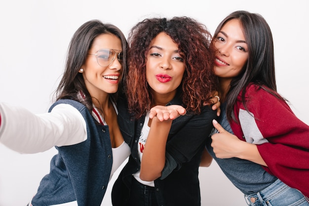 Close-up portrait of charming multiracial girls posing. Elegant african young woman with red lipstick sending air kiss while her friends laughing.
