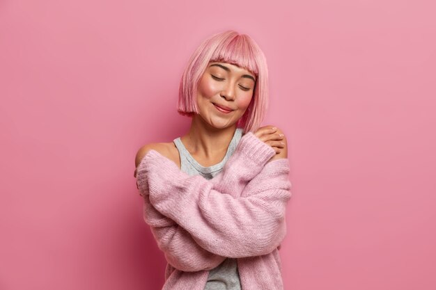 Крупным планом портрет очаровательной симпатичной азиатской женщины с розовой прической, обнимает себя, стоит с закрытыми глазами, носит теплый мягкий свитер, стоит в помещении