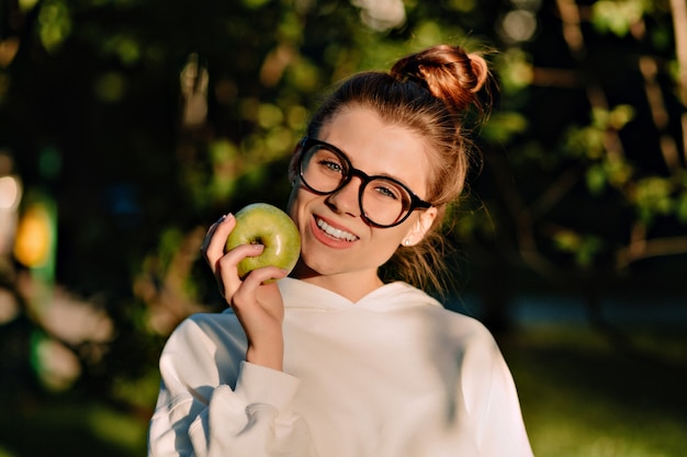 Free photo close up portrait of charming pretty lady wearing spectaculars eating apple in sunlight in the park