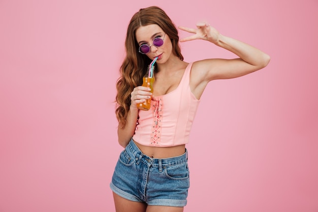 Free photo close-up portrait of charming readhead woman in glasses, drinking orange soda, showing peace sign