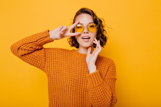 Close-up portrait of charming young woman with gently smile posing with peace sign in trendy glasses