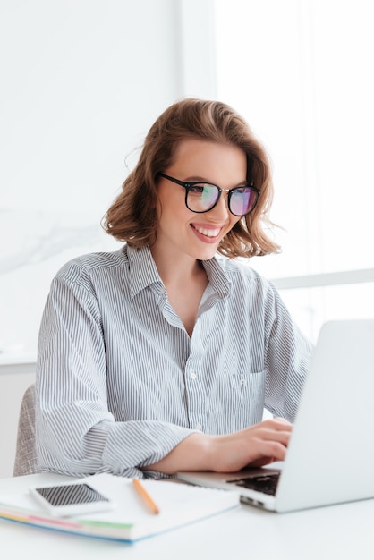 Close-up portrait of cheerful bunette woman in glasses using laptop computer while working at home