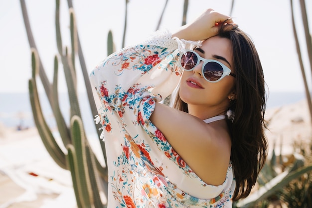 Close-up portrait of cheerful girl in white sunglasses posing with hands up on cactus. Amazing joyful young woman in attire with floral print having fun outside in summer vacation