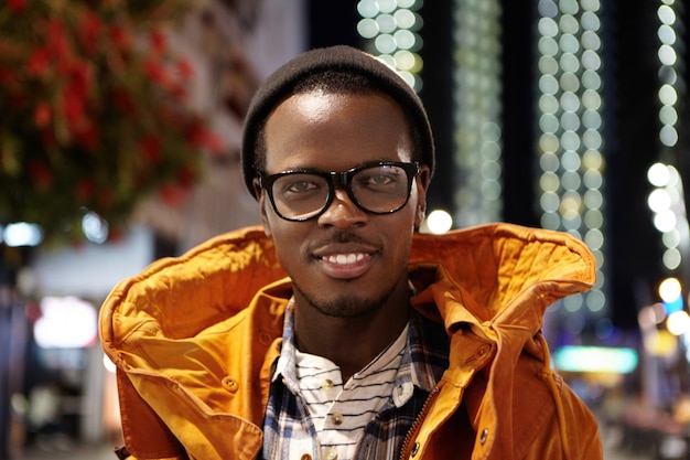 Free photo close up portrait of cheerful and happy young dark-skinned male tourist wearing winter coat, glasses and hat standing on streets of modern metropolis at night, waiting for taxi outside airport