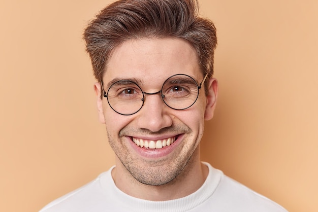 Free photo close up portrait of cheerful man smiles happily shows white teeth wears round spectacles for vision correction feels satisfied isolated over beige background glad to meet with beutiful woman