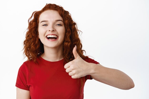close up portrait of cheerful and positive ginger girl with curly long hair, showing thumb up in approval and say yes, recommend good thing, praise product, white wall