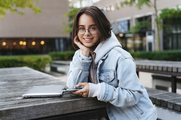 Close-up portrait of cheerful pretty young female student with short hair, lean on palm looking cute at camera with happy smile, sitting near computer, use laptop and mobile phone outdoors.