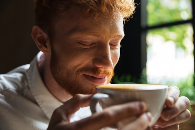 Close-up portrait of cheerful redhead bearded man sniffs coffee with closed eyes