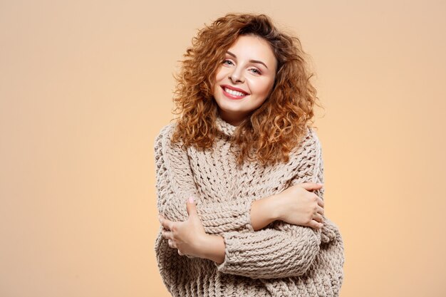 Close up portrait of cheerful smiling beautiful brunette curly girl in knitted sweater over beige wall