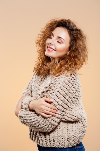 Close up portrait of cheerful smiling beautiful brunette curly girl in knitted sweater over beige wall