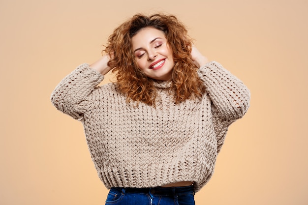 Крупным планом портрет веселой улыбающейся красивой брюнетки кудрявой девушки в вязаном свитере на бежевой стене