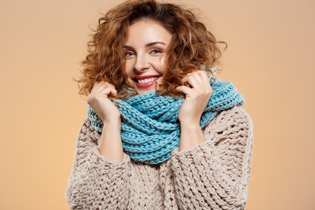 Free photo close up portrait of cheerful smiling beautiful brunette curly girl in knitted sweater and grey neckwarmer over beige wall