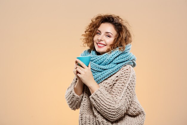 Close up portrait of cheerful smiling beautiful brunette curly girl in knitted sweater and grey neckwarmer holding cup over beige wall