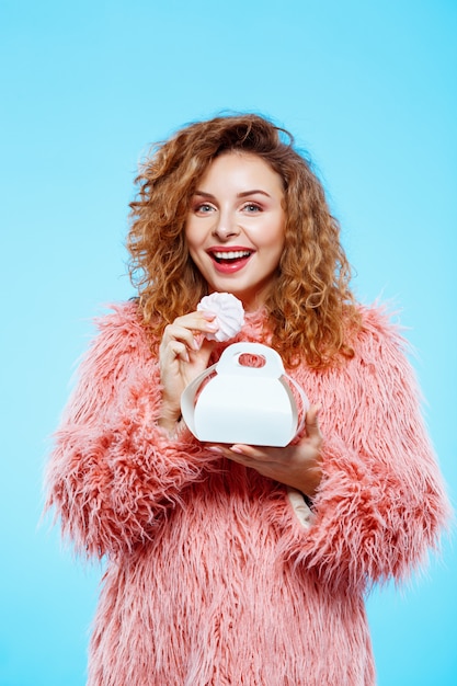 Close up portrait of cheerful smiling beautiful brunette curly girl in pink fur coat eating marshmallow over blue wall