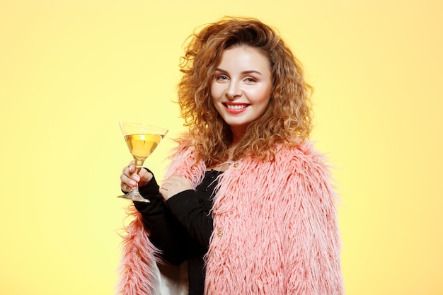 Close up portrait of cheerful smiling beautiful brunette curly girl in pink fur coat holding cocktail glass over yellow wall