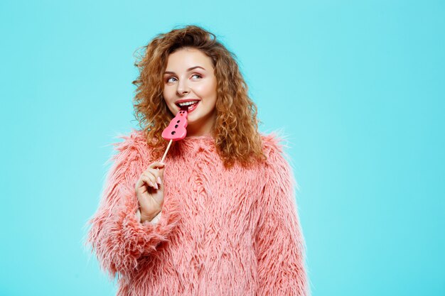 Крупным планом портрет веселой улыбающейся красивой брюнетки кудрявой девушки в розовом меховом пальто леденца на палочке над голубой стеной
