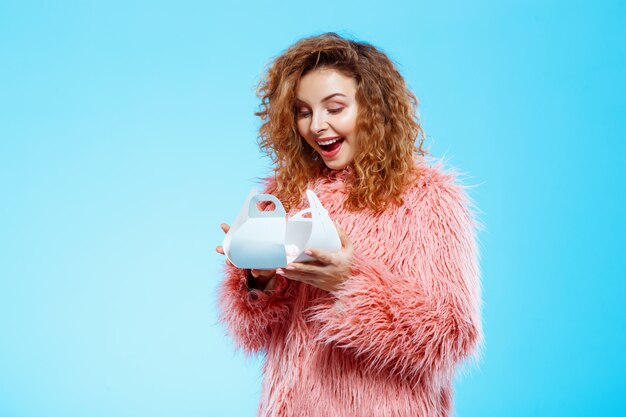 Close up portrait of cheerful smiling surprised beautiful brunette curly girl in pink fur coat over blue wall