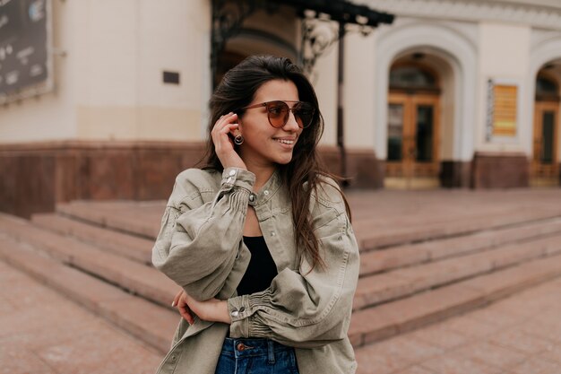 Close up portrait of a cheerful woman with glasses touching her hair in the street