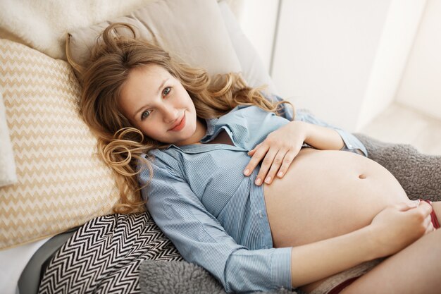 Close up portrait of cheerful young beautiful pregnant woman in home clothes with curly light hair laying in bed with happy and relaxed expression, enjoying motherhood moments.