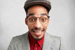 Free photo close up portrait of clumsy comic male wears big spectacles, cap and jacket, smiles with surprisment,