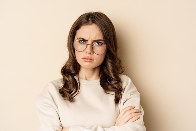 Close up portrait of complicated, troubled woman in glasses, frowning and looking displeased, standing over beige background. Copy space