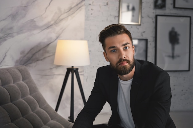 Close up ritratto di fiducioso attraente alla moda giovane imprenditore con barba rifilata e acconciatura alla moda in posa in interni moderni ed accoglienti, vestito in abito elegante formale,