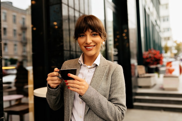 Close up portrait of confident smiling modern business woman with a cup of coffee against the backdrop of a large business building High quality photo