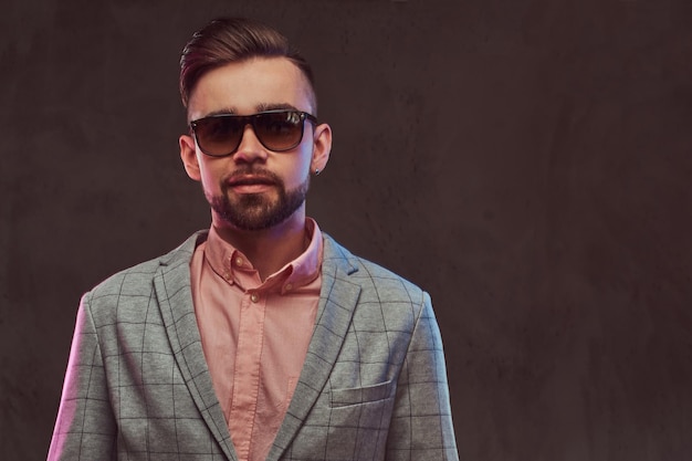 Close-up portrait of a confident stylish bearded man with hairstyle and sunglasses in a gray suit and pink shirt, posing in a studio. Isolated on a gray background.