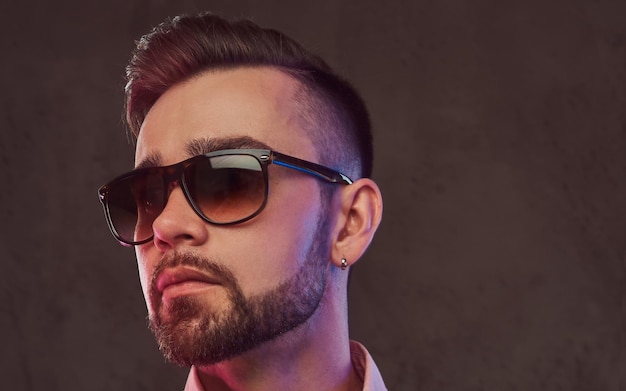 Close-up portrait of a confident stylish bearded man with hairstyle and sunglasses in a gray suit and pink shirt, posing in a studio. Isolated on a gray background.