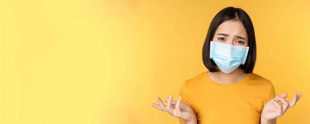 Close up portrait of confused asian woman in medical face mask shrugging shoulders and looking puzzl