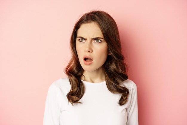 Close up portrait of confused, frustrated young woman, looking left with disappointment, puzzled by something, standing over pink background