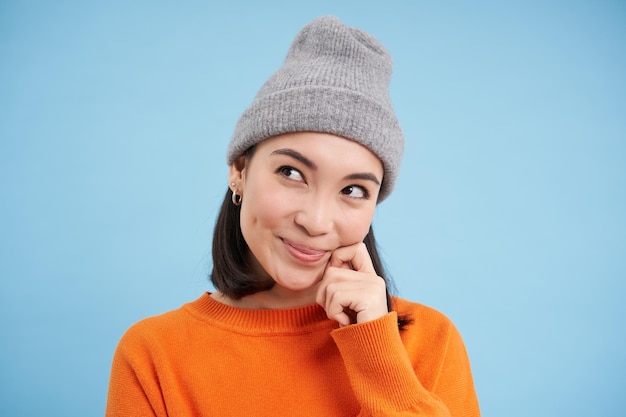 Free photo close up portrait of coquettish asian girl in hat looks aside and smiling fantasizing dreaming of sm