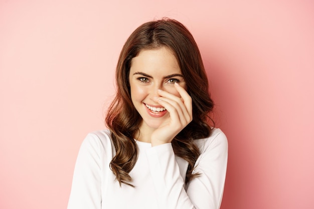 Free photo close up portrait of coquettish brunette woman, laughing and smiling, looking down flirty, standing over pink background