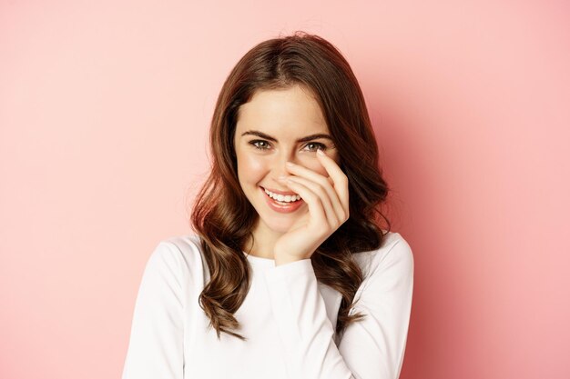 Close up portrait of coquettish brunette woman, laughing and smiling, looking down flirty, standing over pink background