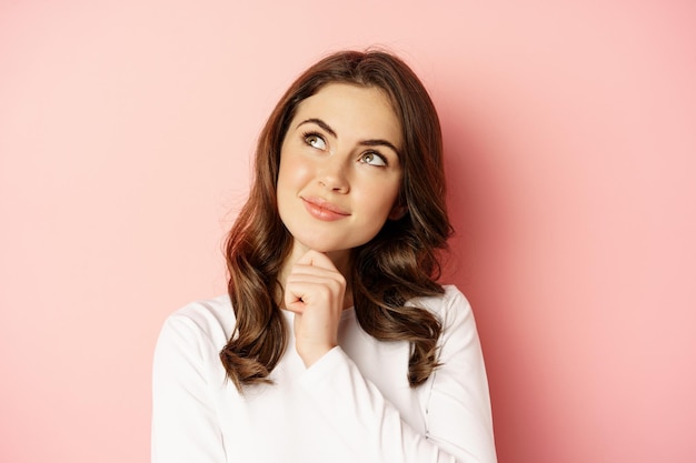 Close up portrait of coquettish smiling woman, glamour girl thinking, looking thoughtful, standing over pink background