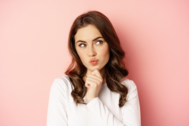 Close up portrait of coquettish smiling woman, glamour girl thinking, looking thoughtful, standing over pink background.