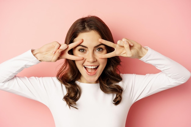 Free photo close up portrait of coquettish young woman smiling showing peace vsign gesture and posing happy sta...