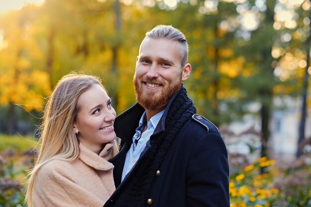 Close up portrait of a couple on a date walk in autumn park. Positive redhead bearded male hugs the cute blonde female.