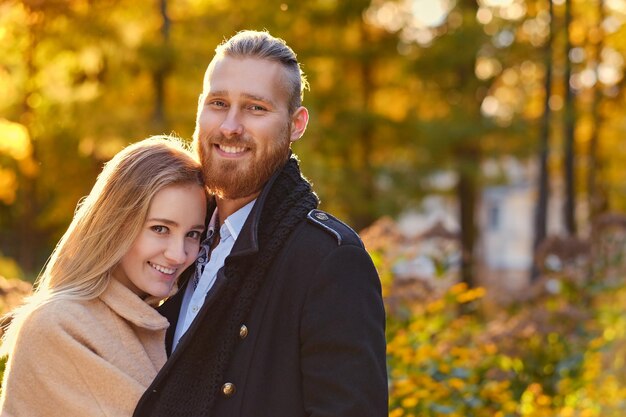Close up portrait of a couple on a date walk in autumn park. Positive redhead bearded male hugs the cute blonde female.
