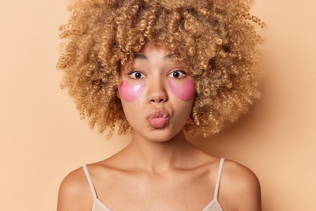 Close up portrait of curly haired woman keeps lips folded wants to kiss someone applies pink hydrogel patches under eyes for rejuvenation wears casual t shirt isolated over beige background.